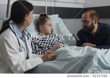 Pediatrician examining ill kid health condition while discussing with parent. Caring father sitting next to sick daughter resting in hospital pediatric ward while doctor presenting treatment scheme. Pediatrician examining ill kid health condition while discussing with parent. Caring father sitting next to sick daughter resting in hospital pediatric ward while doctor presenting treatment scheme. 92147160