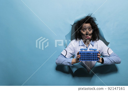Crazy chemist with funny look and wacky hair sticking tongue out while having rack filled with plastic test tubes. Mad scientist with dirty face and goofy expression having stand filled with samples. Crazy chemist with funny look and wacky hair sticking tongue out while having rack filled with plastic test tubes. Mad scientist with dirty face and goofy expression having stand filled with samples. 92147161