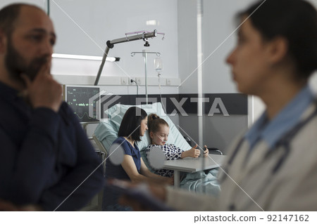 Sick girl playing games on smartphone with mother while father talking with expert pediatrist. Hospitalized ill little kid watching cartoons on mobile phone while resting in hospital pediatric ward 92147162