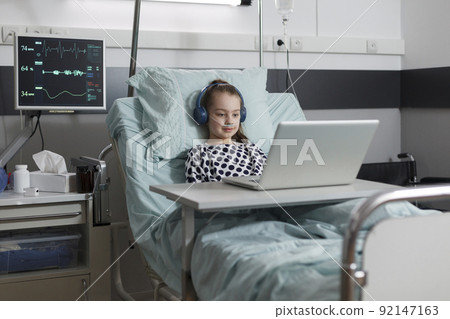 Young sick patient under treatment watching cartoons on laptop while resting on patient bed inside healthcare facility pediatrics ward room. Ill little girl with computer enjoying internet content Young sick patient under treatment watching cartoons on laptop while resting on patient bed inside healthcare facility pediatrics ward room. Ill little girl with computer enjoying internet content 92147163