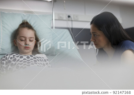Uneasy parent sitting beside sick daughter hospitalized in pediatrics ward. Ill little girl resting inside healthcare facility treatment room while mother sitting next to her. Uneasy parent sitting beside sick daughter hospitalized in pediatrics ward. Ill little girl resting inside healthcare facility treatment room while mother sitting next to her. 92147168