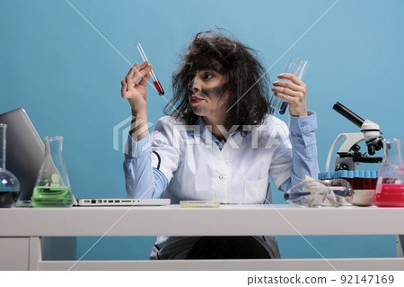 Mad scientist with goofy aspect in laboratory examinating liquid substance. Foolish wild looking crazy chemist analyzing glass test tubes filled with blood sample while sitting at desk in laboratory. Mad scientist with goofy aspect in laboratory examinating liquid substance. Foolish wild looking crazy chemist analyzing glass test tubes filled with blood sample while sitting at desk in laboratory. 92147169