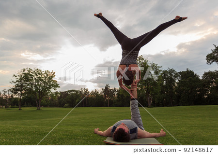 Man lying on grass and balancing woman in his feet. Young couple doing acro yoga in park Man lying on grass and balancing woman in his feet. Young couple doing acro yoga in park 92148617