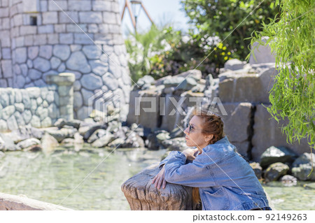 A woman sits in the shade of trees, leaning on a wooden railing against the background of a stone castle and water. Peaceful relaxation in the amusement park. Selective focus. A woman sits in the shade of trees, leaning on a wooden railing against the background of a stone castle and water. Peaceful relaxation in the amusement park. Selective focus. 92149263