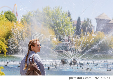 A woman stands in front of a fountain gushing from a pond in an amusement park on a sunny day 92149264