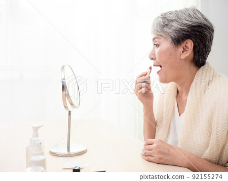 Portrait of mature aged asian woman 60s applying lipstick looking at mirror. Portrait of mature aged asian woman 60s applying lipstick looking at mirror. 92155274