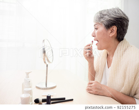 Portrait of mature aged asian woman 60s applying lipstick looking at mirror. Portrait of mature aged asian woman 60s applying lipstick looking at mirror. 92155275