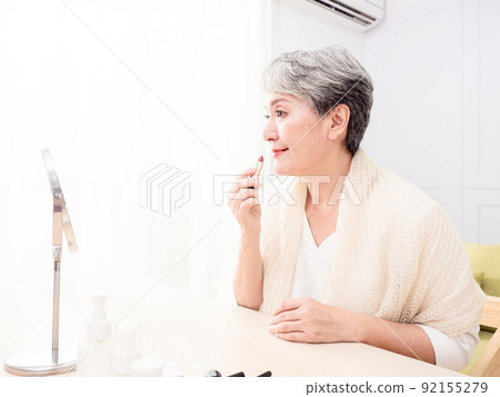 Portrait of mature aged asian woman 60s applying lipstick looking at mirror. Portrait of mature aged asian woman 60s applying lipstick looking at mirror. 92155279