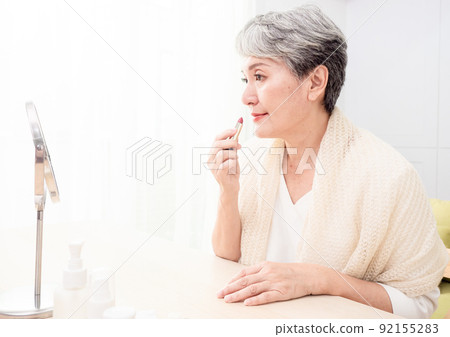 Portrait of mature aged asian woman 60s applying lipstick looking at mirror. 92155283