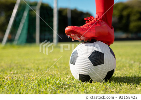 Low section of male athlete wearing red socks and shoes putting leg on soccer ball over grassy land 92158422