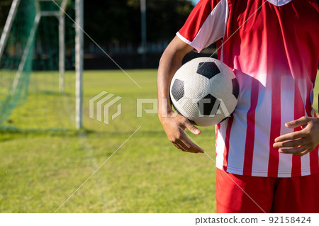 Midsection of caucasian male player wearing red jersey with soccer ball standing at playground 92158424