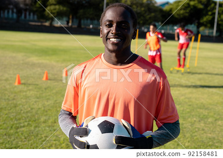 Portrait of african american smiling goalkeeper wearing gloves holding soccer ball at playground Portrait of african american smiling goalkeeper wearing gloves holding soccer ball at playground 92158481