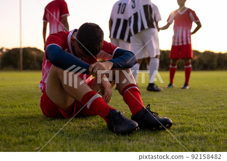 Caucasian sad male athlete sitting on grassy land with team players in background at playground 92158482