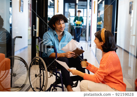 Businesswoman discussing with young businessman sitting in wheelchair at office corridor 92159028