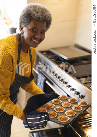 Vertical image of happy african american senior woman taking out cupcakes from oven 92159394