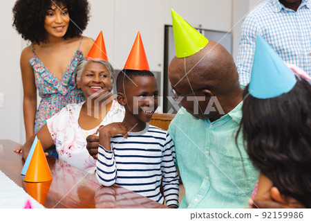 Happy multiracial multigeneration family wearing colorful party hats while enjoying birthday at home 92159986