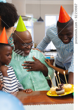 Multiracial senior man wearing party hat looking at cupcakes while celebrating birthday with family 92159987
