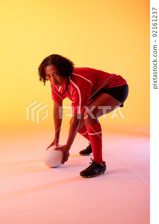 Portrait of african american female rugby player with rugby ball over neon yellow lighting 92161237
