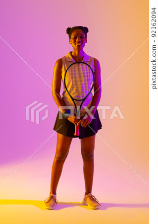 Vertical image of happy african american female tennis player in neon violet and yellow lighting Vertical image of happy african american female tennis player in neon violet and yellow lighting 92162094