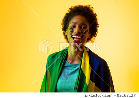 Image of african american female soccer fan with flag of brazil cheering in yellow lighting 92162127