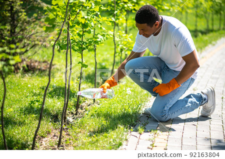 Man crouched watering small tree in park Man crouched watering small tree in park 92163804