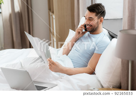 man with folder calling on phone in bed at home 92165033