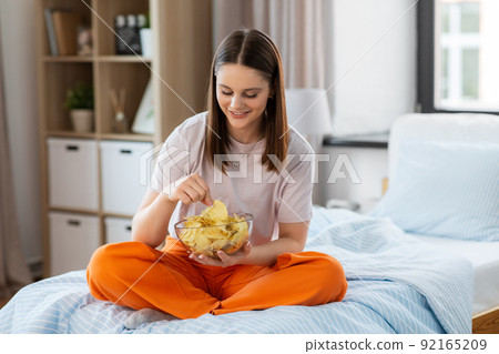 happy girl eating crisps sitting on bed at home 92165209