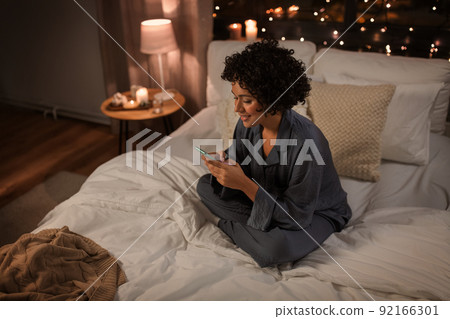 happy woman in pajamas with phone in bed at night happy woman in pajamas with phone in bed at night 92166301
