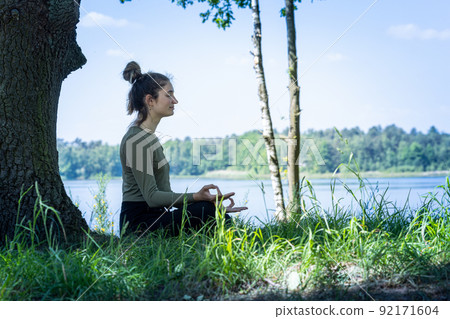 Young woman practicing yoga in nature. Sitting on green grass next to a tree and a forest lake 92171604