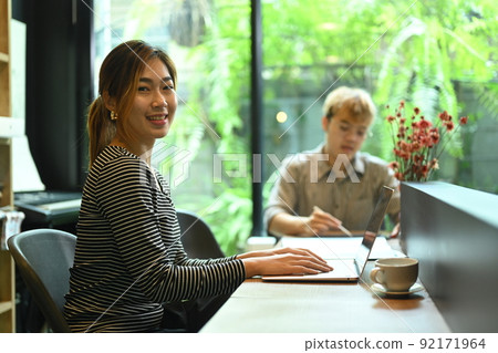 Confident female website developer sitting in modern office with her colleague and smiling to camera Confident female website developer sitting in modern office with her colleague and smiling to camera 92171964