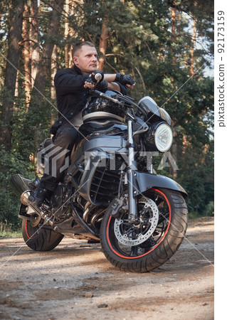 Young male biker travels on a motorcycle alone, stopped on the side of a forest road Young male biker travels on a motorcycle alone, stopped on the side of a forest road 92173159
