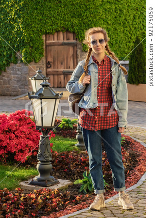 A woman in a modern street outfit walks near a flower bed with bright pink flowers and low decorative lanterns against a stone house overgrown with foliage. Weekend in the park 92175059