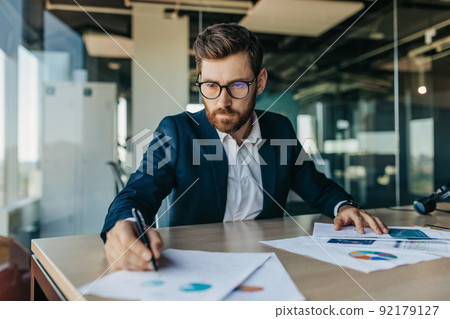 Concentrated handsome businessman in glasses working with documents in office interior, checking and signing papers 92179127