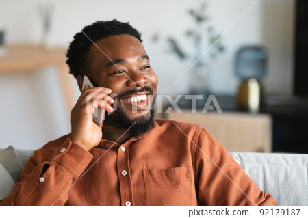 Cheerful Black Guy Talking On Cellphone Sitting On Sofa Indoor 92179187