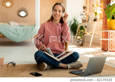 Happy teenage female student in headphones reading book during online class near laptop computer, sitting at home Happy teenage female student in headphones reading book during online class near laptop computer, sitting at home 92179326