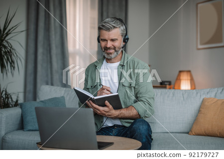 Smiling happy aged european man with beard in headphones has meeting remotely at computer 92179727