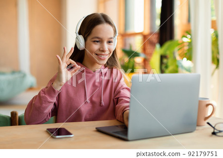 Studentgirl having online video class via laptop and gesturing OK, approving e-learning, sitting at desk and smiling 92179751