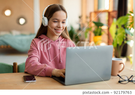 Kids and technology concept. Happy teen girl sitting at table and using laptop, wearing wireless headphones, copy space 92179763