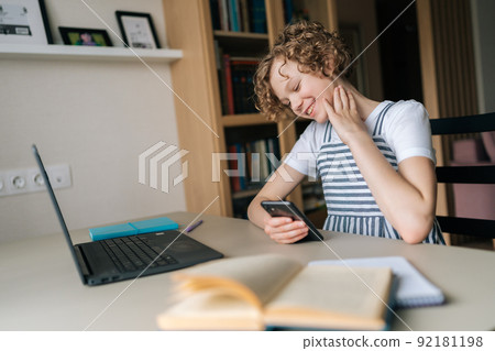 Positive adorable little girl holding and using mobile phone, browsing social network apps, surfing internet and scrolling news feed, reading electronic e-book, sitting at table with laptop and books. 92181198