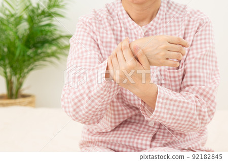 Middle-aged woman wearing pajamas and holding her wrist in the bedroom Middle-aged woman wearing pajamas and holding her wrist in the bedroom 92182845