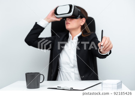 Female Executive Holding Pen And Learning Professional Skill Through Virtual Reality Simulator. Woman Wearing Suit Sitting At Desk And Experiencing Modern Technology. Female Executive Holding Pen And Learning Professional Skill Through Virtual Reality Simulator. Woman Wearing Suit Sitting At Desk And Experiencing Modern Technology. 92185426