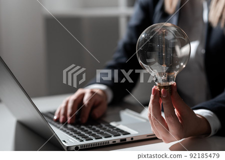 Businesswoman Typing Recent Updates On Lap Top On Desk Holding Lightbulb. Woman In Office Writing Important Message On Computer. Executive Inserting Crutial Data Into Pc. 92185479