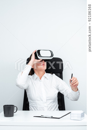 Businesswoman Wearing Virtual Reality Goggles And Learning Professional Skill Through Futuristic Simulation. Woman Holding Pen And Sitting At Desk During Modern Training. Businesswoman Wearing Virtual Reality Goggles And Learning Professional Skill Through Futuristic Simulation. Woman Holding Pen And Sitting At Desk During Modern Training. 92186168