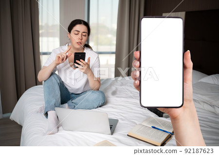 A young caucasian woman sits on the bed and uses a smartphone. Close-up of a hand holding a smartphone. Mock up. The concept of online education and dependence of social networks 92187623