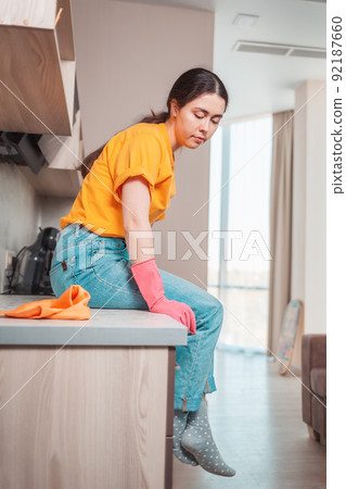 A housewife in casual clothing and rubber gloves sits on the kitchen table, resting after cleaning. Side view 92187660