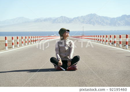 asian woman tourist sitting in the middle of an empty open road looking at view with lake and rolling mountains in background, leg crossed 92195460