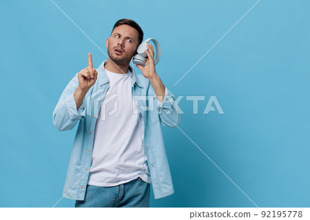 Thoughtful pensive tanned handsome man in casual basic t-shirt hold listen headphones look point finger up posing isolated on blue studio background. Copy space Banner Mockup. Music concept 92195778