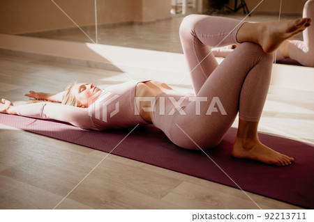 Young sporty woman, fitness instructor in pink sportswear doing stretching and pilates on yoga mat in the studio with mirror. Female fitness yoga routine concept. Healthy lifestyle and harmony. Young sporty woman, fitness instructor in pink sportswear doing stretching and pilates on yoga mat in the studio with mirror. Female fitness yoga routine concept. Healthy lifestyle and harmony. 92213711