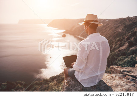 Digital nomad, freelancer. Remote work on vacation. Man in the hat, a businessman with a laptop sits on the rocks by the sea during sunset, makes a business transaction online from a distance. Digital nomad, freelancer. Remote work on vacation. Man in the hat, a businessman with a laptop sits on the rocks by the sea during sunset, makes a business transaction online from a distance. 92213917