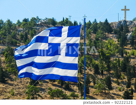 The national flag of Greece on the flagpole develops in the wind against the background of a hill with green trees and a cross on top. Greek island of Kalymnos. 92222121